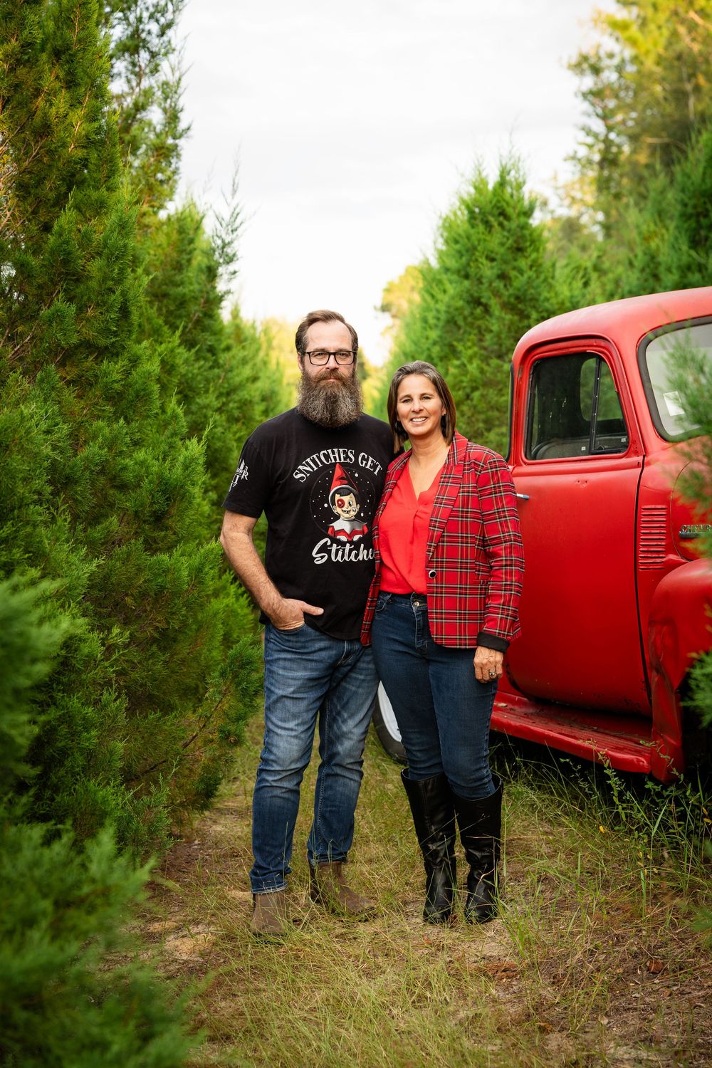 A couple poses together in front of a vintage red pickup truck surrounded by evergreen trees in an outdoor setting.