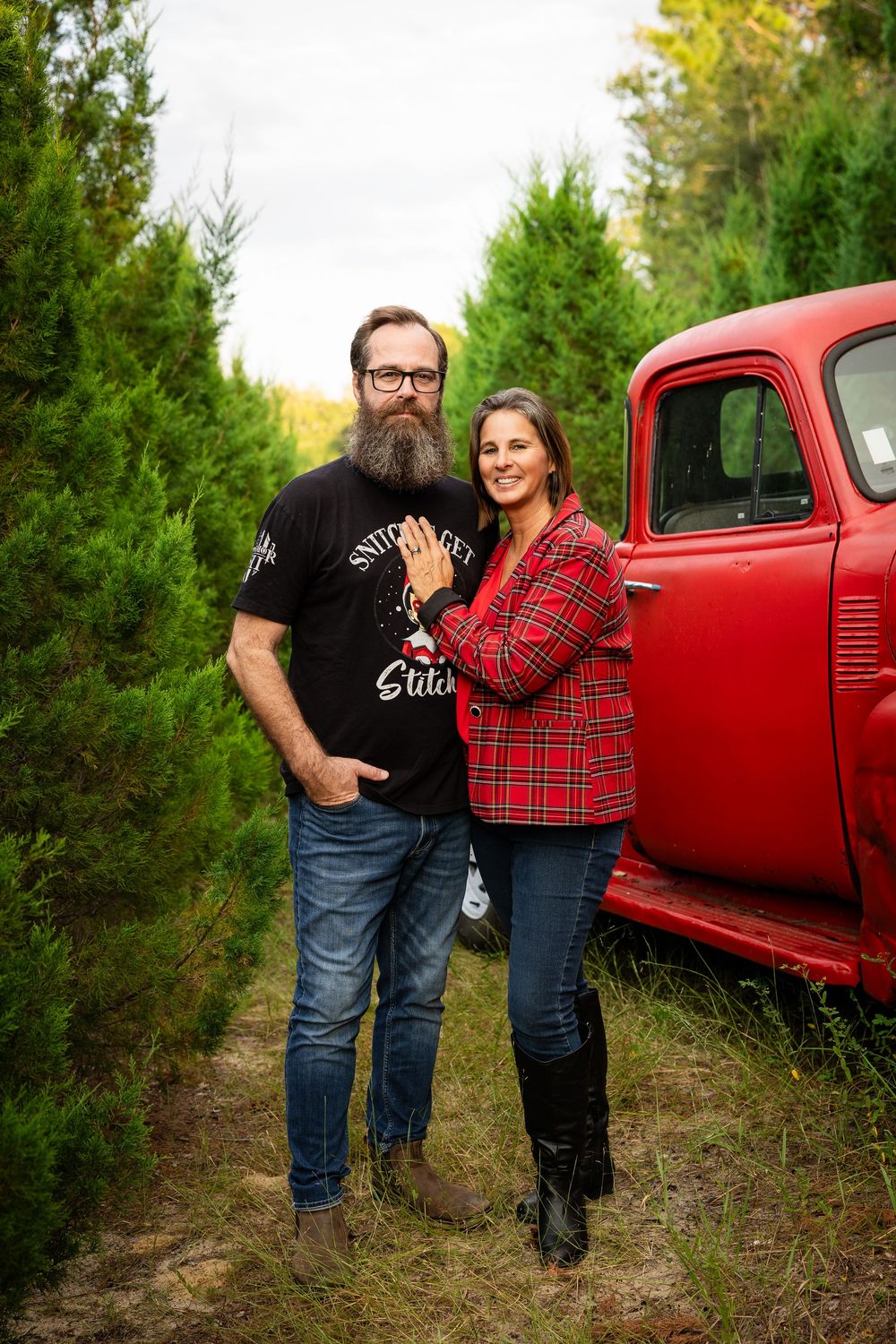 A couple poses together in front of a red vintage truck surrounded by evergreen trees for outdoor photos.