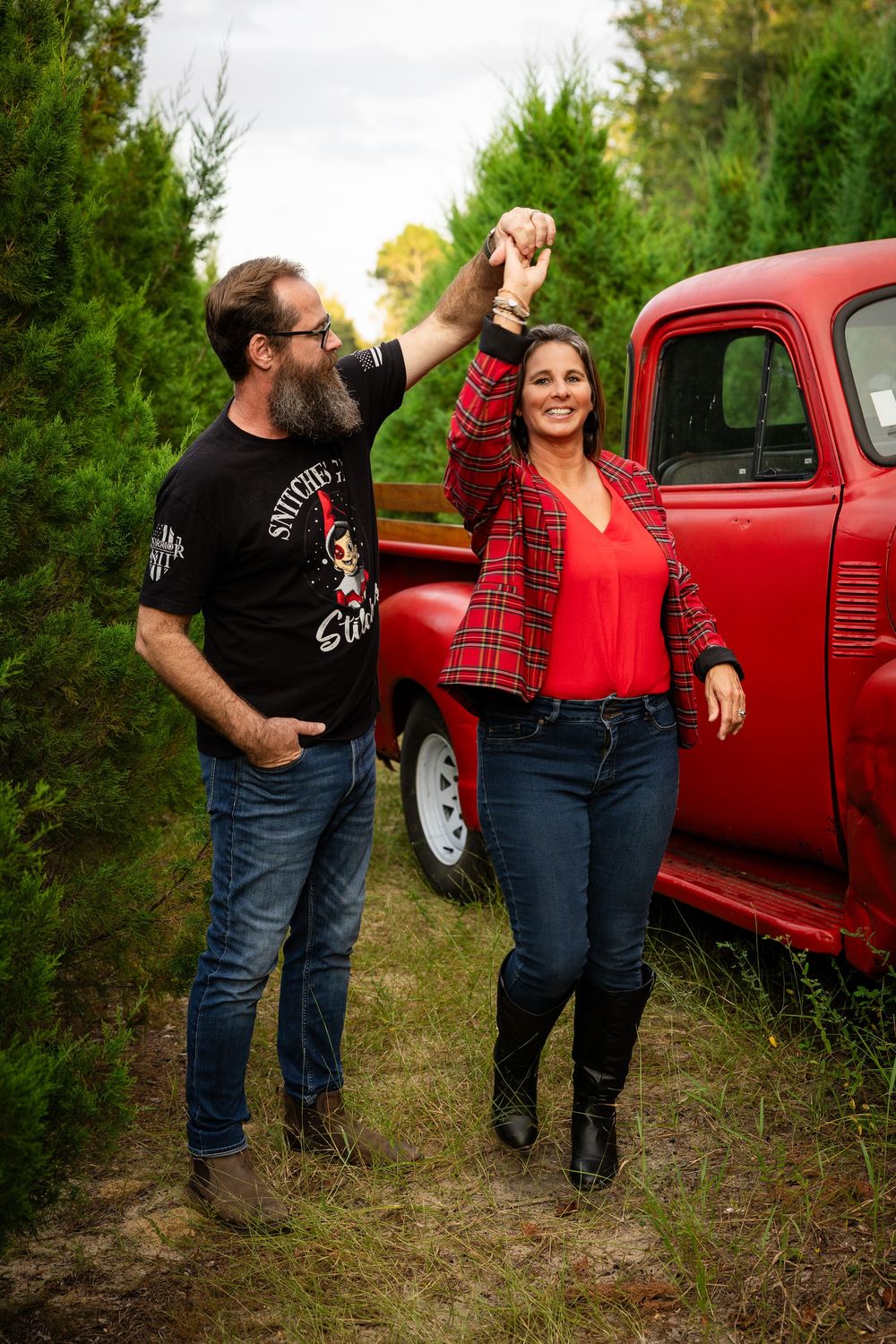 A couple poses playfully in front of a vintage red pickup truck in a wooded outdoor setting.