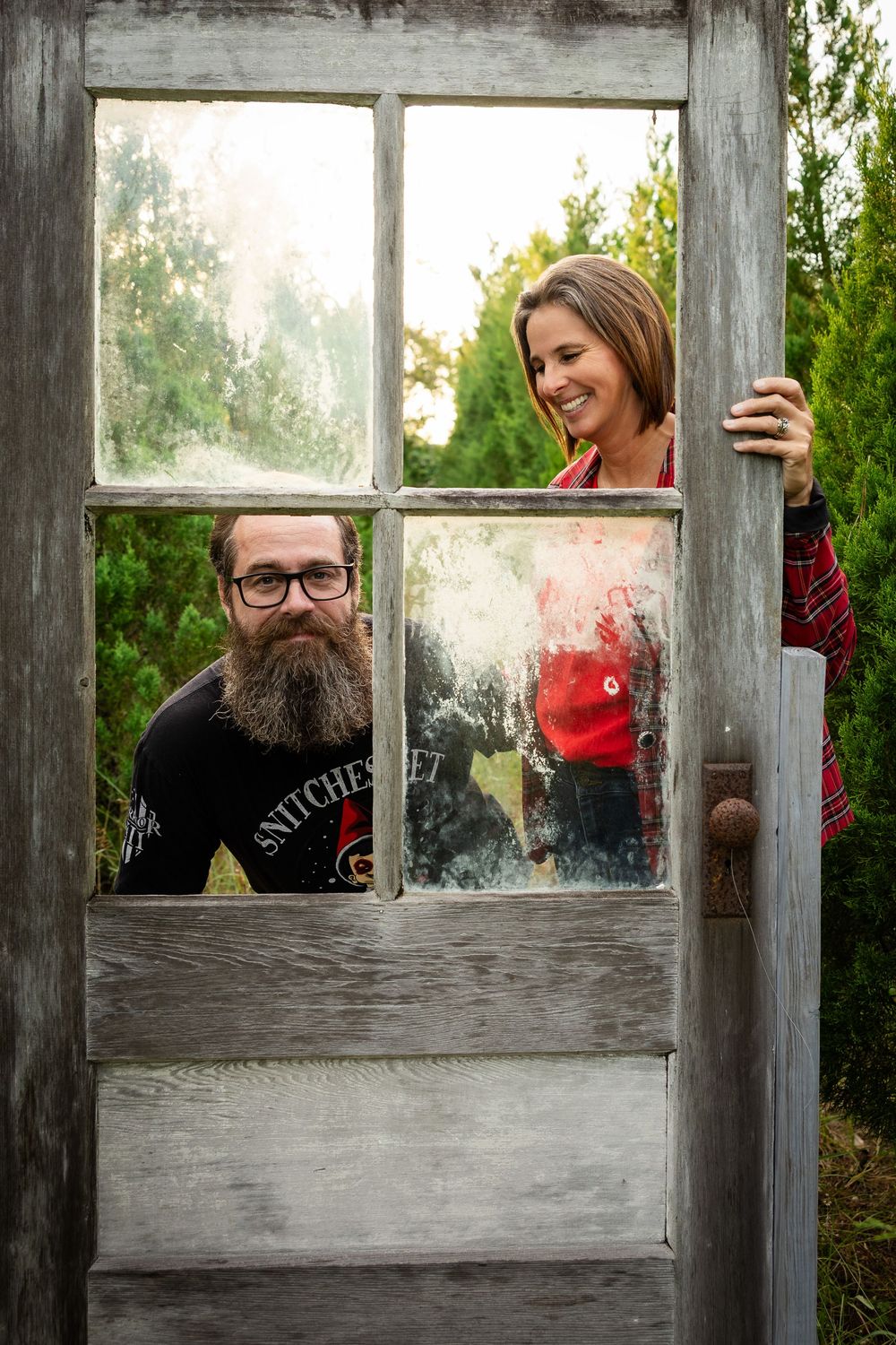 Two people peek playfully through the panes of a weathered wooden door frame in an outdoor garden setting.