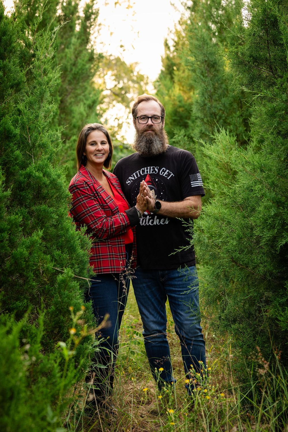 A couple poses together among tall evergreen trees wearing coordinated plaid and black outfits in an outdoor setting.