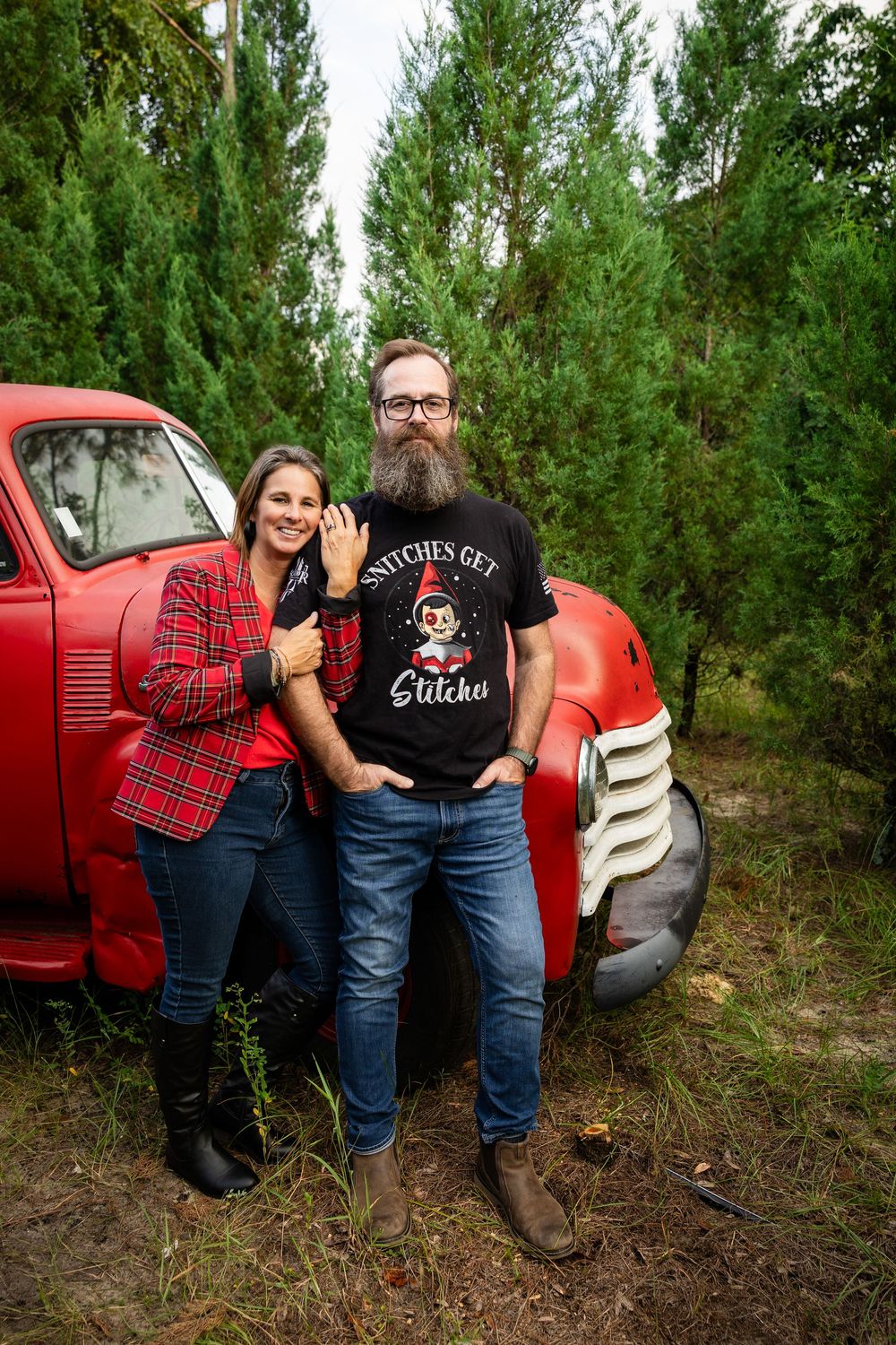 Couple in matching plaid shirts pose casually by a vintage red truck in a pine forest setting.