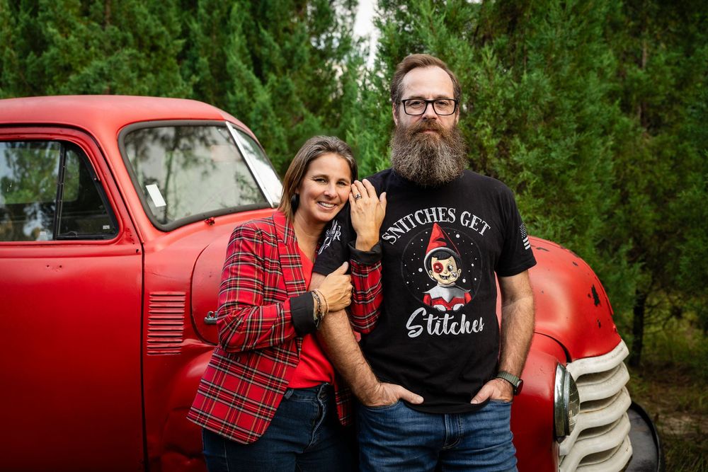 A couple poses in front of a vintage red truck wearing coordinated red plaid and black outfits in a green outdoor setting.