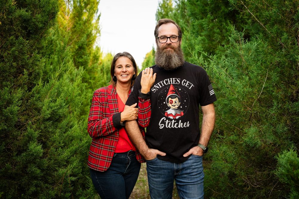 A couple in plaid and black shirts pose together against a lush green forest backdrop.