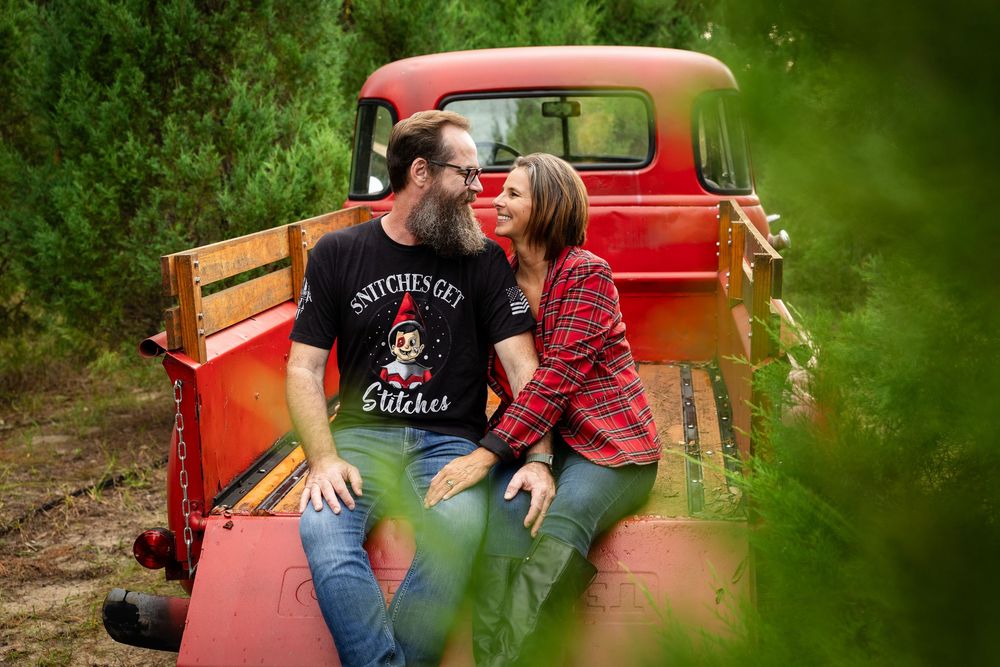 A couple sits together in the bed of a vintage red pickup truck surrounded by lush green foliage.