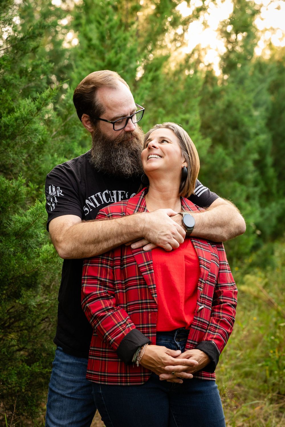 A couple embraces outdoors wearing coordinating red plaid and black outfits against a green natural background.