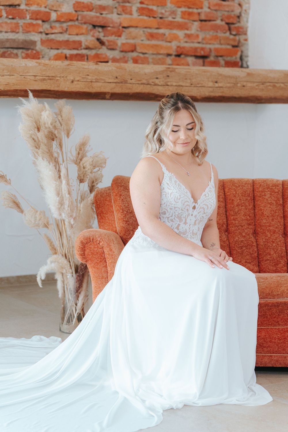 A bride in a white wedding gown sits gracefully on a rustic couch against exposed brick walls with pampas grass decor.