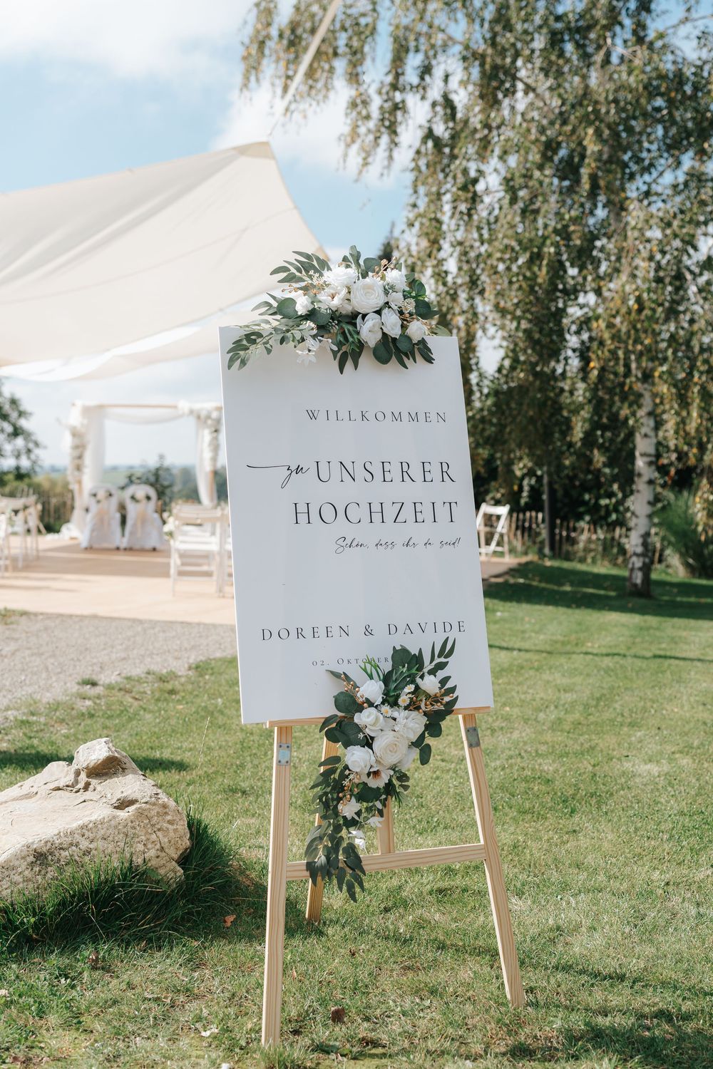 Elegant wedding welcome sign decorated with white flowers and greenery stands on wooden easel at outdoor garden ceremony.
