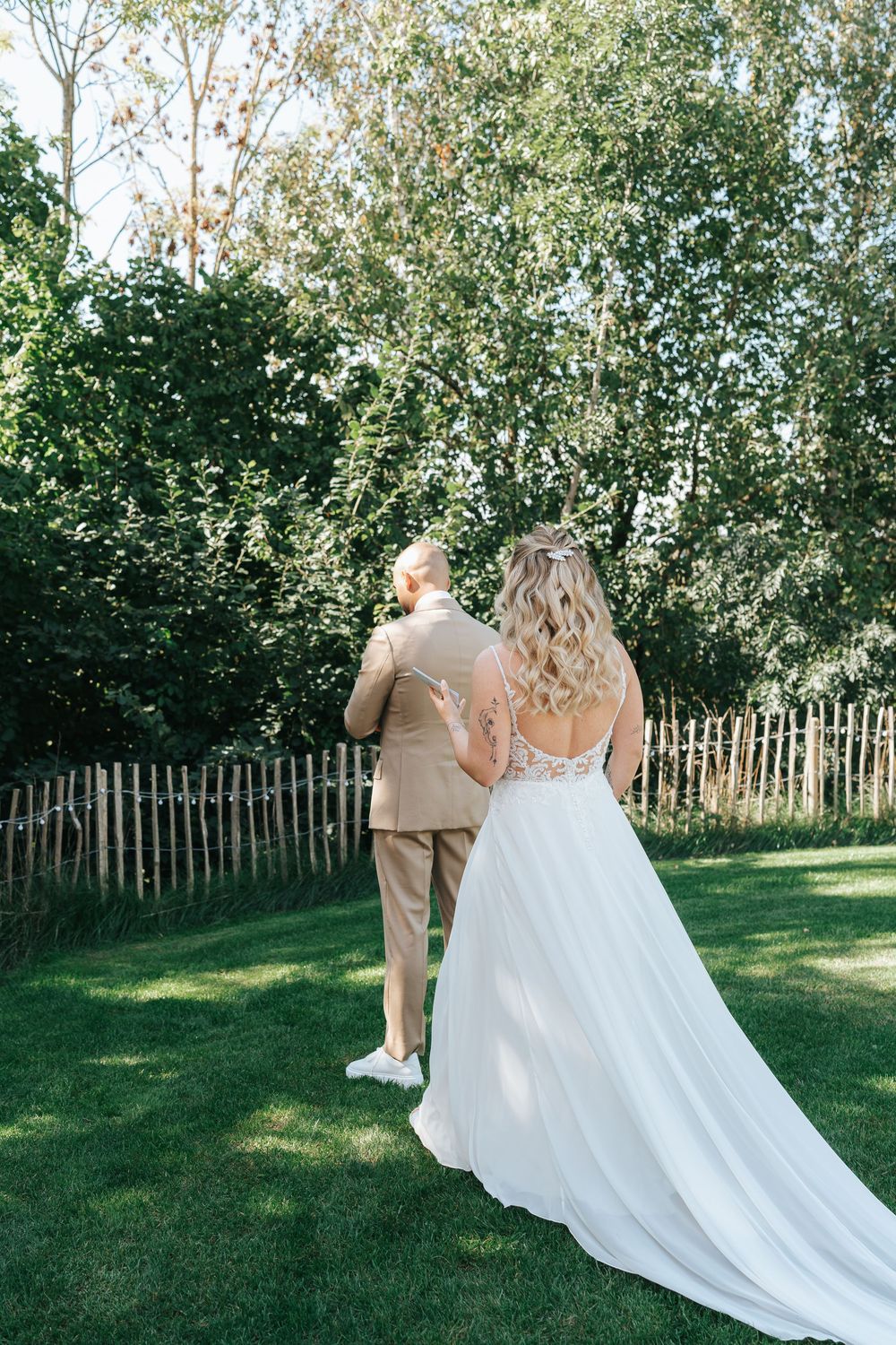 A couple in wedding attire walks together on green grass with a rustic wooden fence and trees in the background.