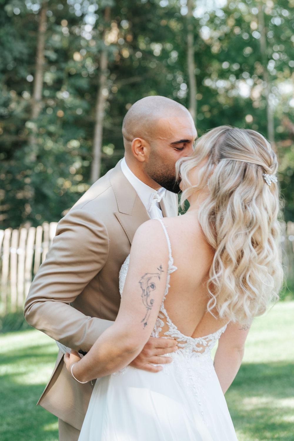 Romantic wedding couple shares tender kisses in a garden setting with trees and sunlight in the background.