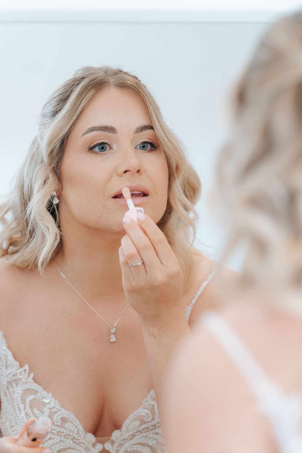 Woman in lace wedding dress applying lip gloss while looking in mirror during bridal preparation.