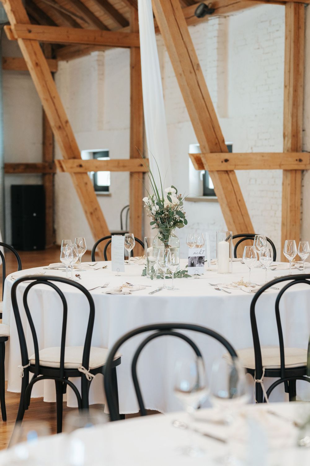 Elegant wedding reception venue with exposed wooden beams, white tablecloths, and black bentwood chairs in a rustic setting.