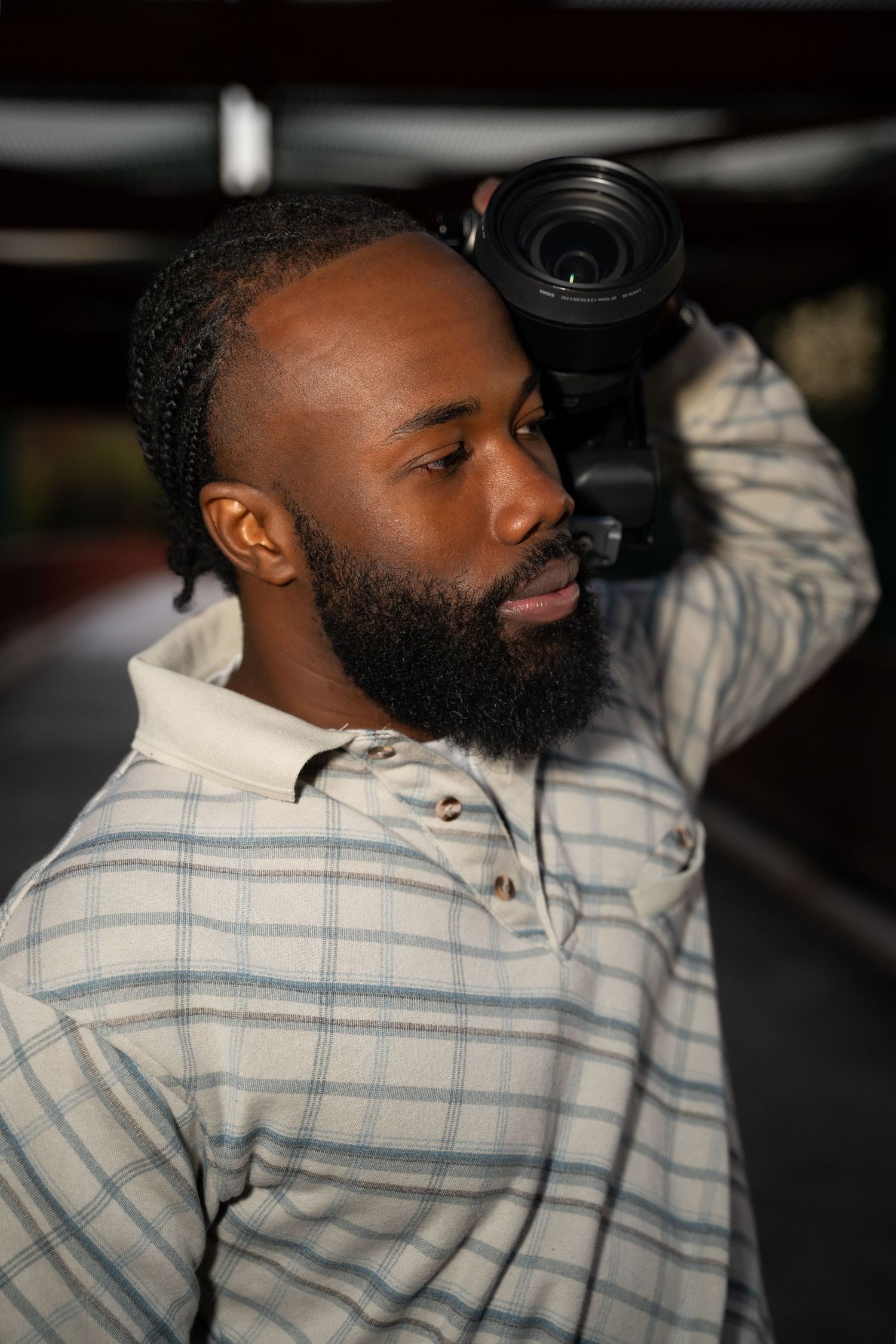 A photographer wearing a plaid shirt holds a camera lens while looking off to the side in a thoughtful pose.