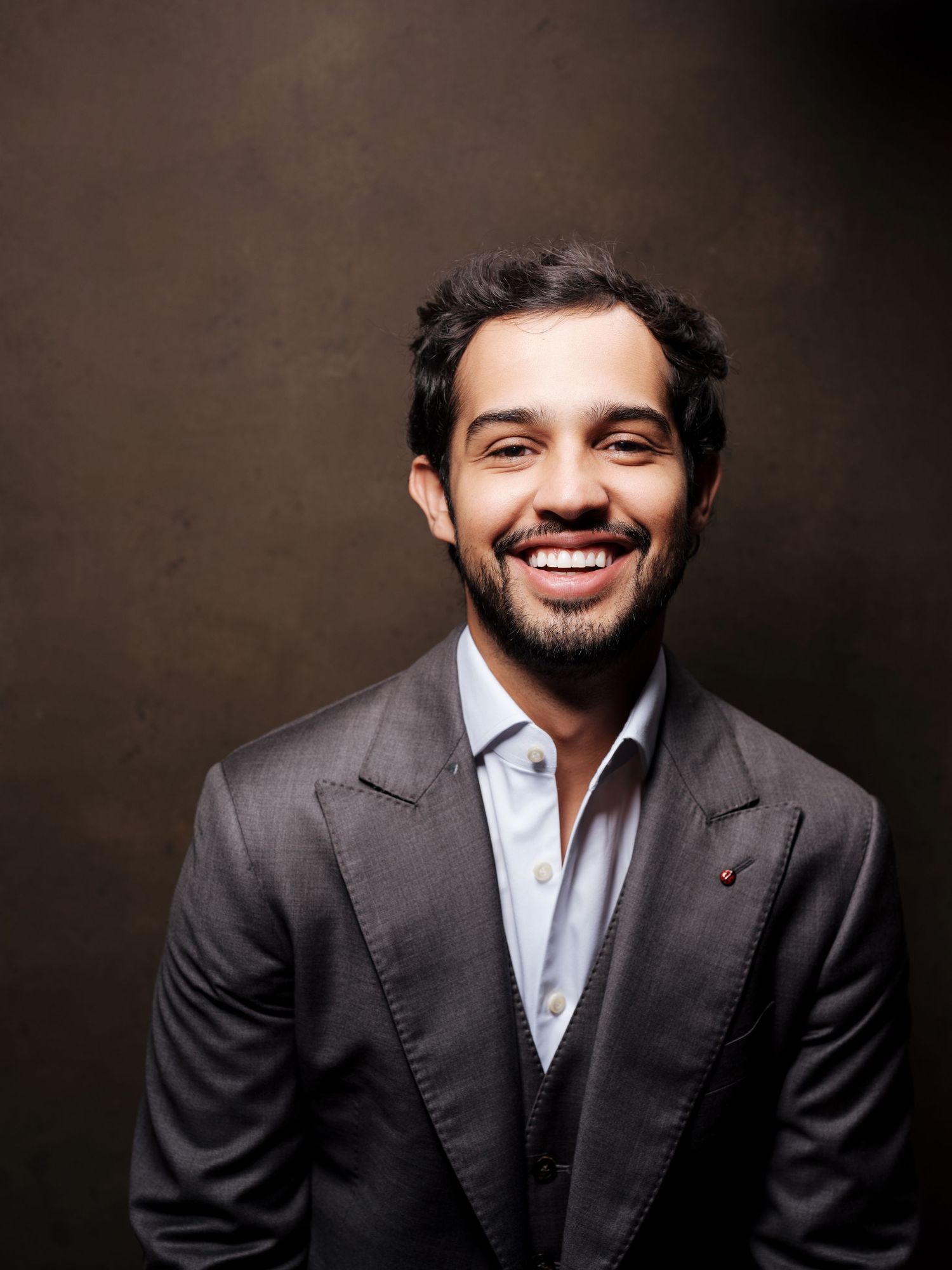 A professional studio portrait of a smiling person wearing a grey suit and white dress shirt against a dark background.