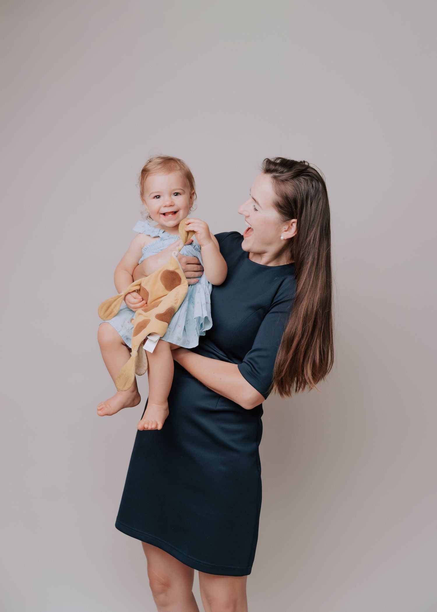 A woman in a black dress holds and plays with a baby wearing light colored clothing against a gray backdrop.