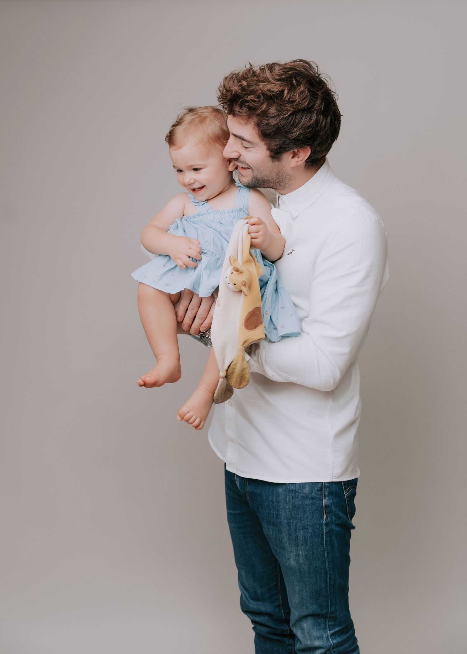 A person in a white shirt and jeans holds a baby in a light blue outfit against a gray background.