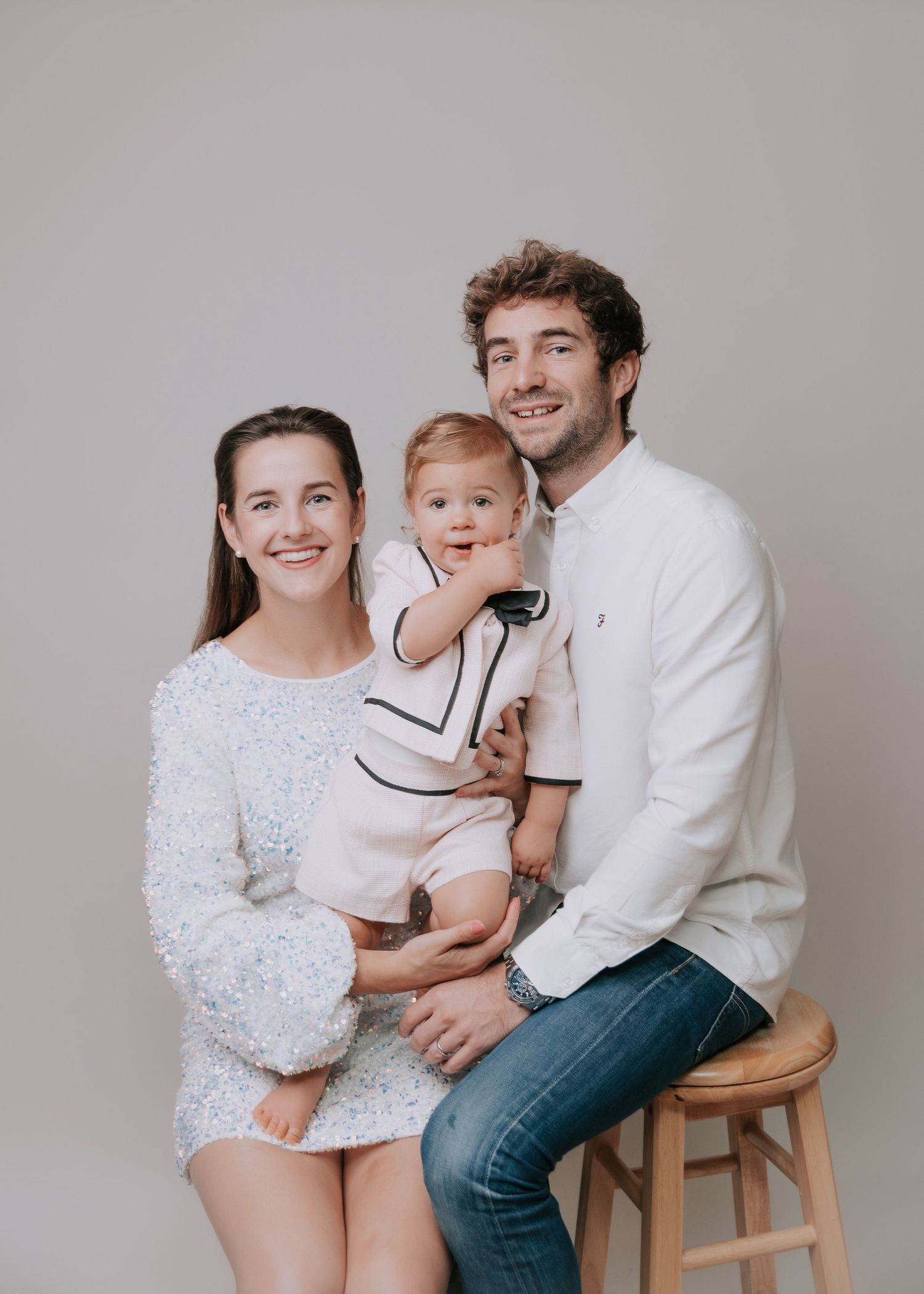 A family wearing white and denim sits together on wooden stools against a neutral backdrop in a studio setting.