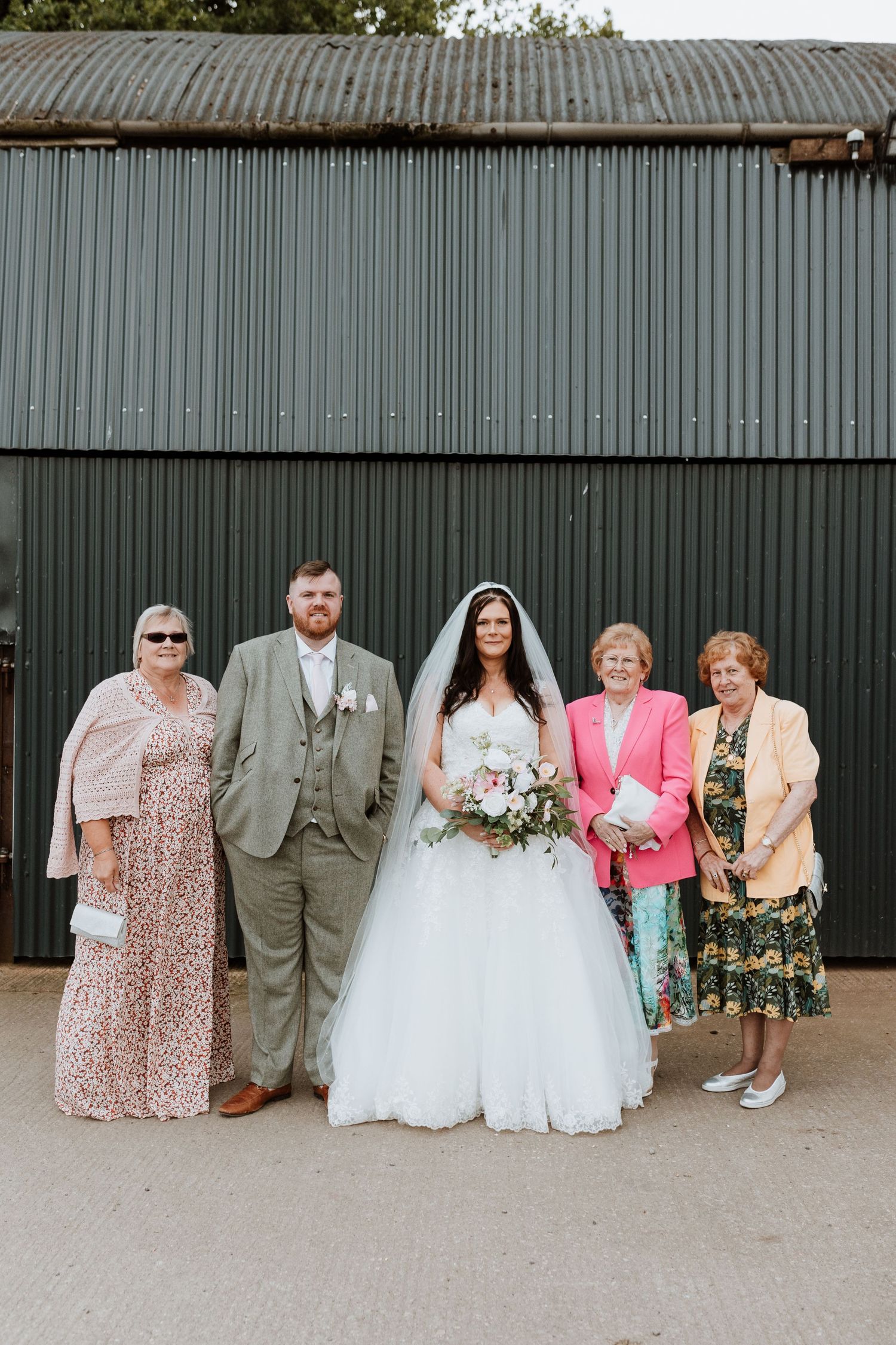 Wedding party sits together on a bench against a rustic metal wall wearing formal attire and a white bridal gown.