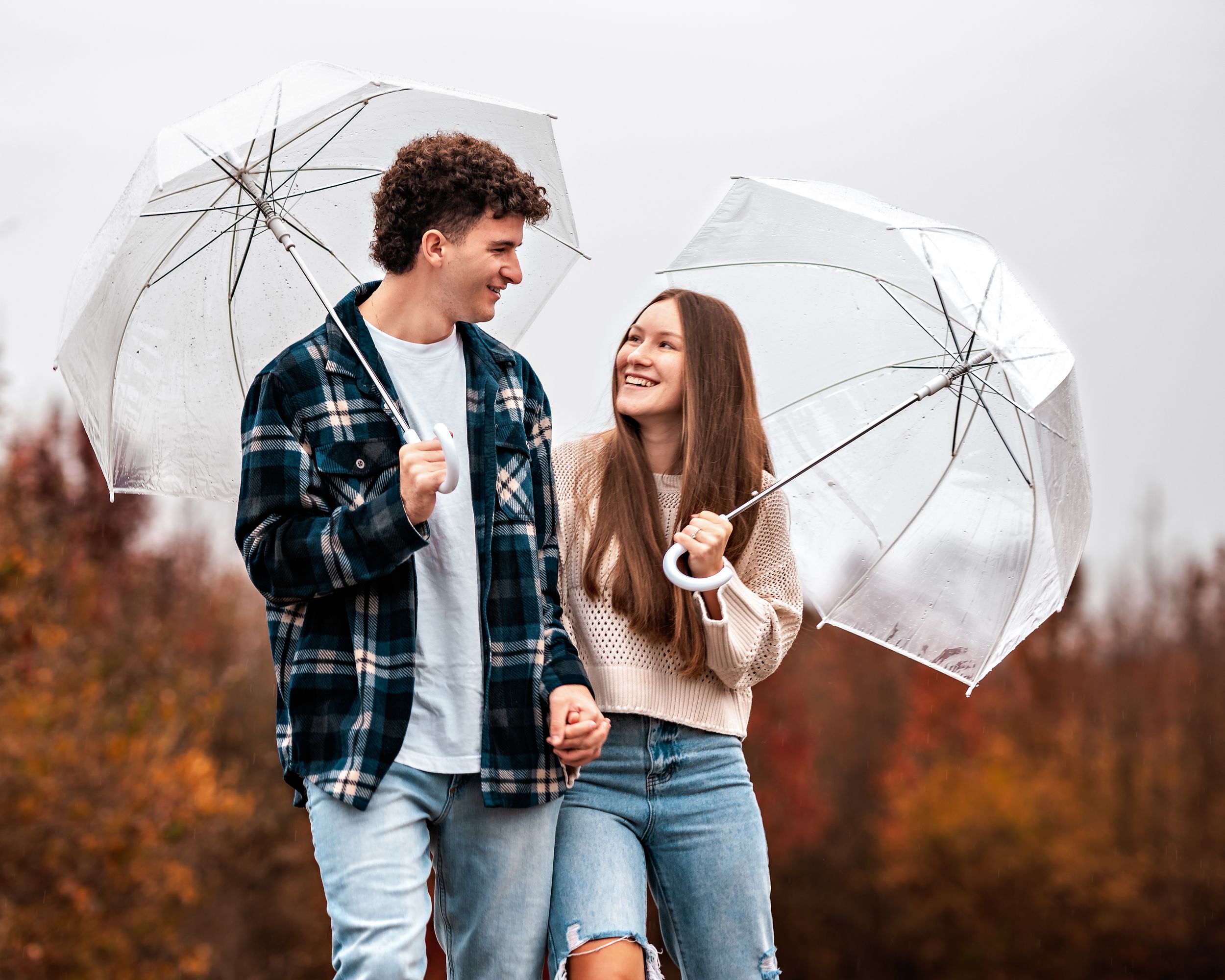 A couple shares a laugh while holding white umbrellas in an autumn setting with fall foliage in the background.