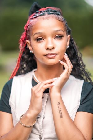 Portrait of a person with braided hair styled with red accents wearing a white vest and black shirt, posing outdoors.