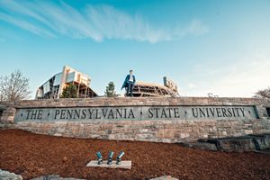 Students sit on the iconic Pennsylvania State University sign wall against a bright blue sky with wispy clouds.