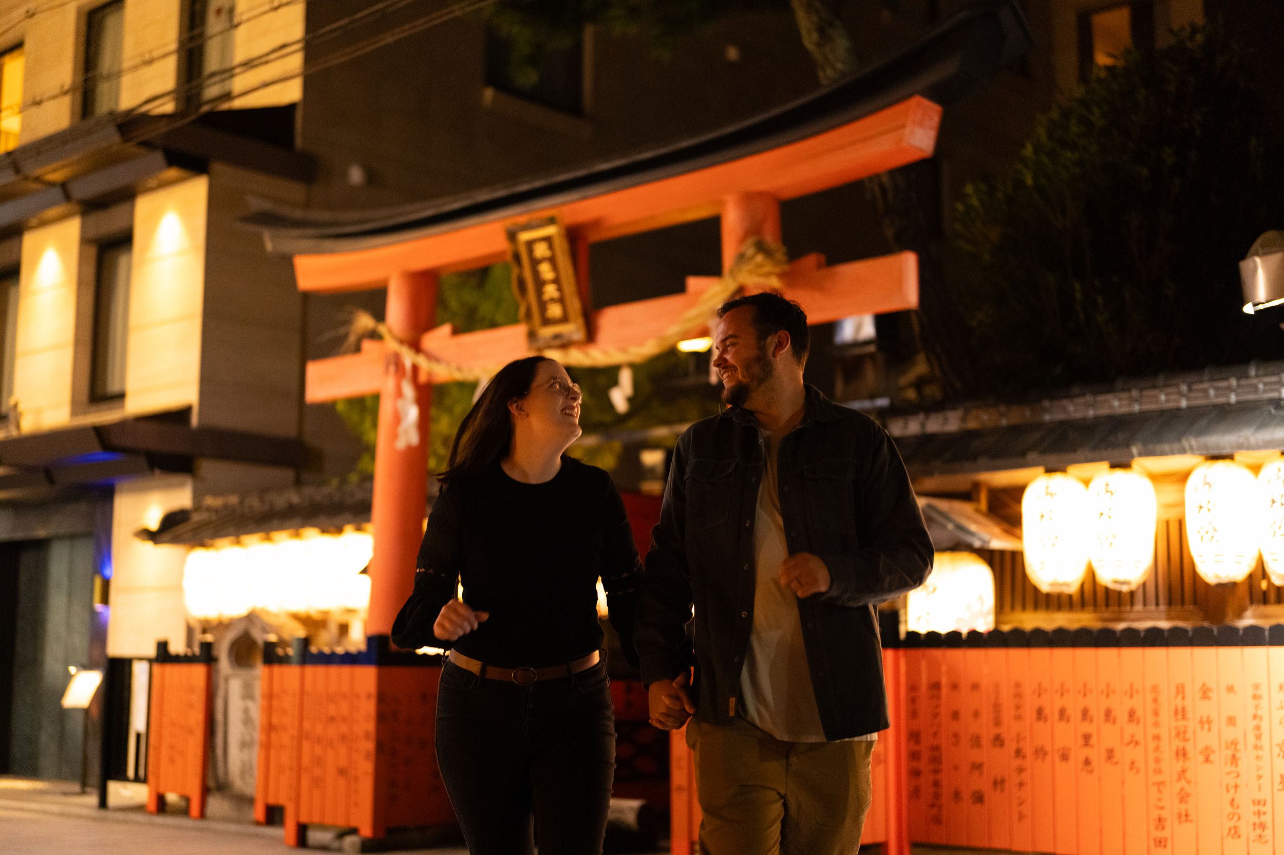 Smiling couple walking near a glowing red torii gate and paper lanterns in Kyoto at night.