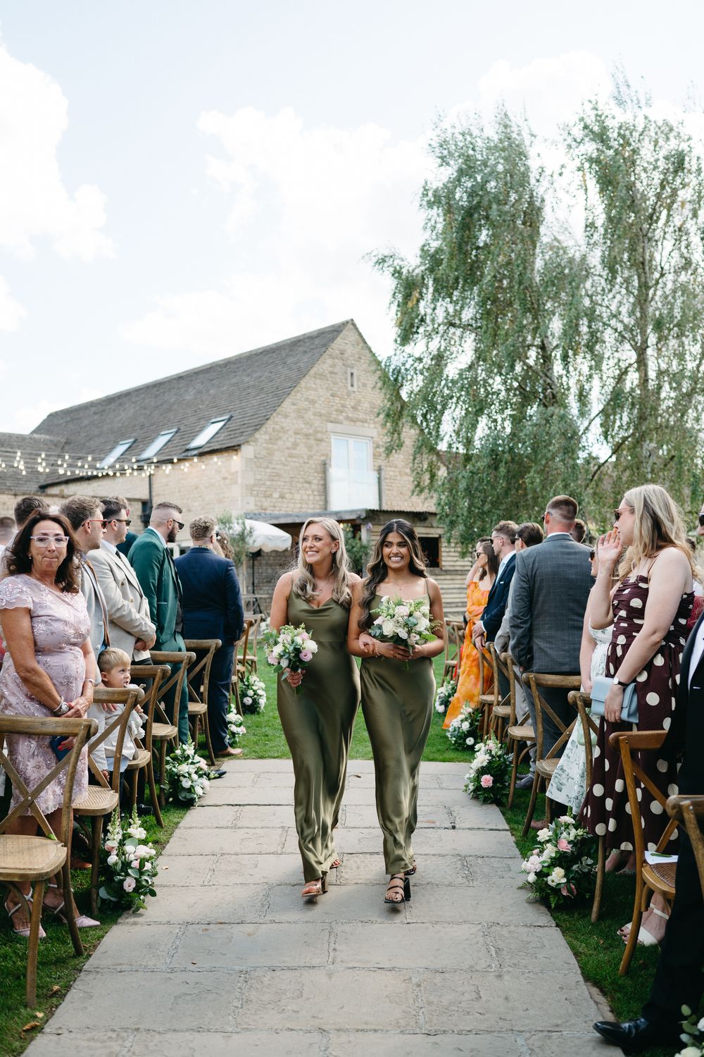 Wedding guests walk down a stone pathway lined with floral arrangements at an outdoor ceremony venue.