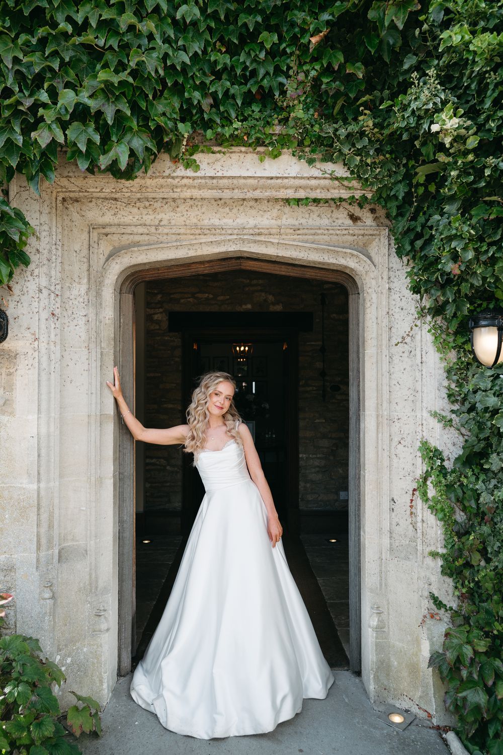 A bride in a white wedding gown poses playfully in a sequence of shots against an ivy-covered stone archway entrance.