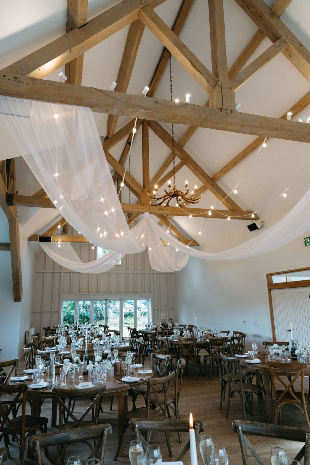 Elegant wedding reception venue with exposed wooden beams, white drapes, and tables set for dining under soft lighting.
