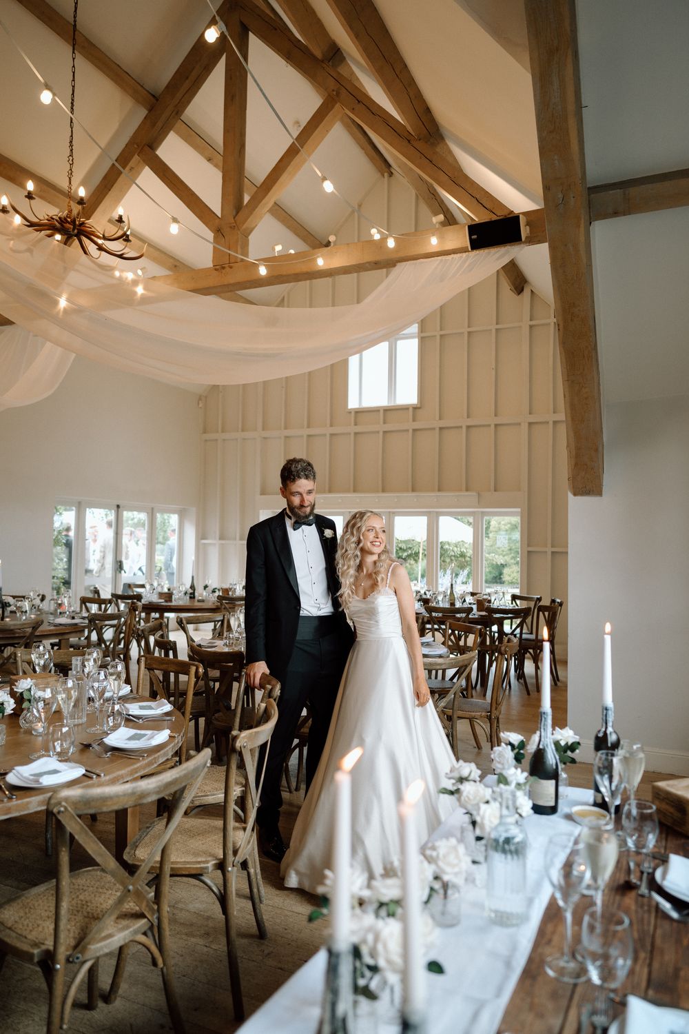 Rustic barn wedding reception venue with exposed wooden beams, elegant table settings and white fabric draping from ceiling.