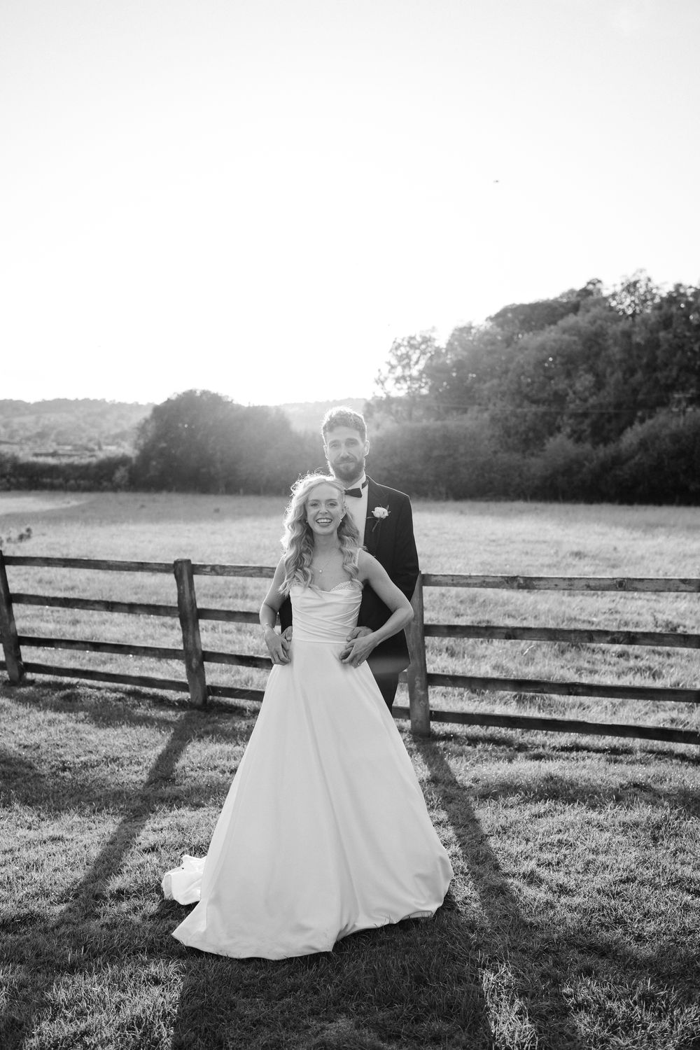 A romantic wedding couple embraces in a sunlit field with rustic wooden fencing at golden hour.