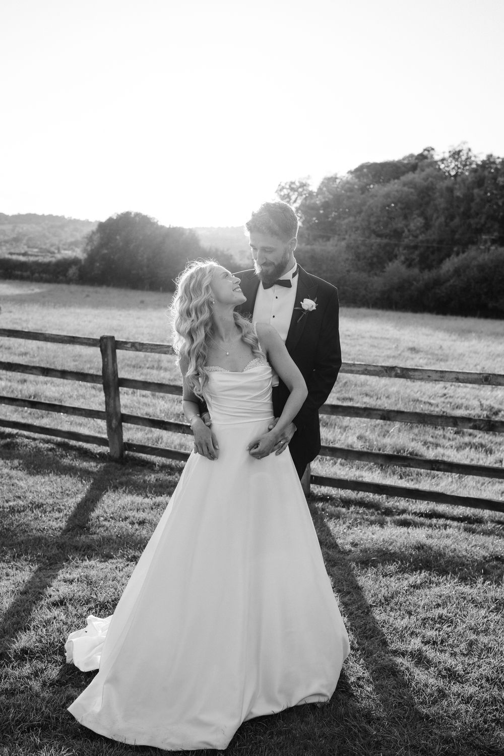 A romantic couple in wedding attire share an intimate moment by a rustic wooden fence at sunset in a countryside setting.
