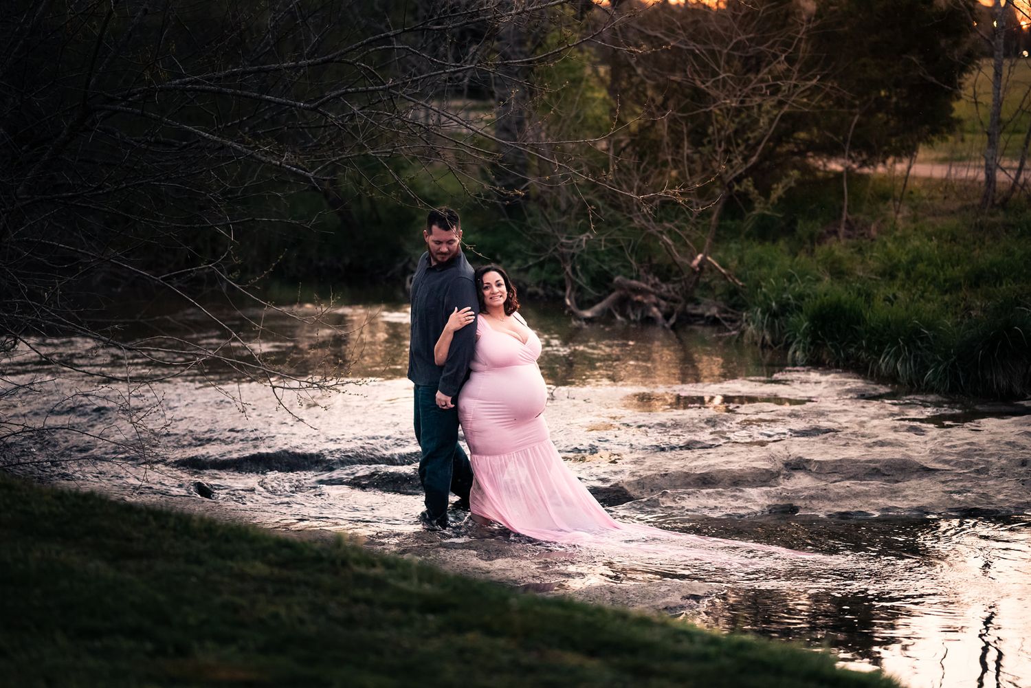 A couple in formal attire poses dramatically at sunset by a lake.