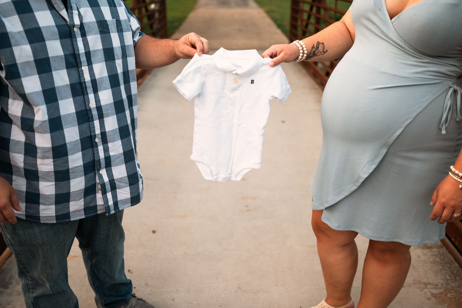 A couple in coordinating blue outfits stands together during a maternity photo shoot in a garden setting.