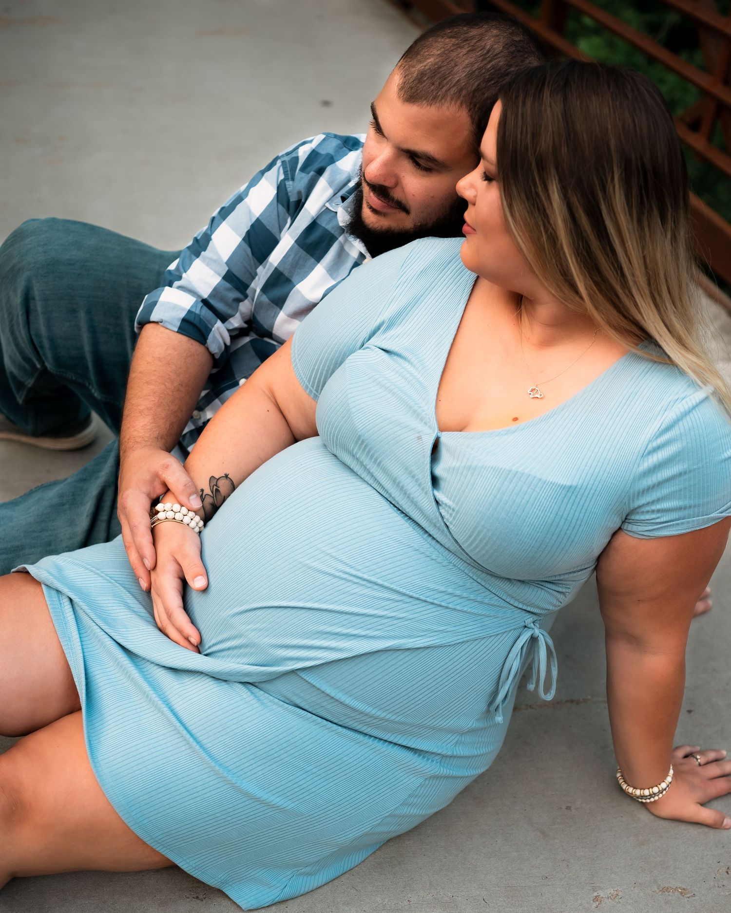 A couple in casual attire sits together intimately on the ground, with the woman wearing a light blue dress.