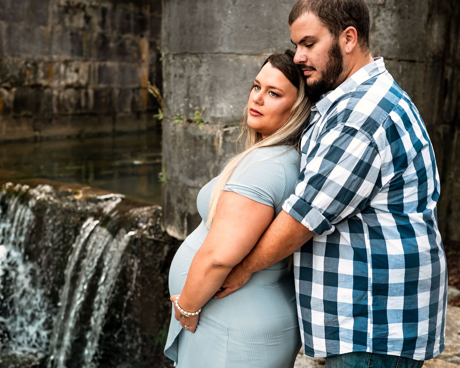 Couple in plaid shirt and gray dress embrace lovingly against stone wall backdrop with cascading waterfall.
