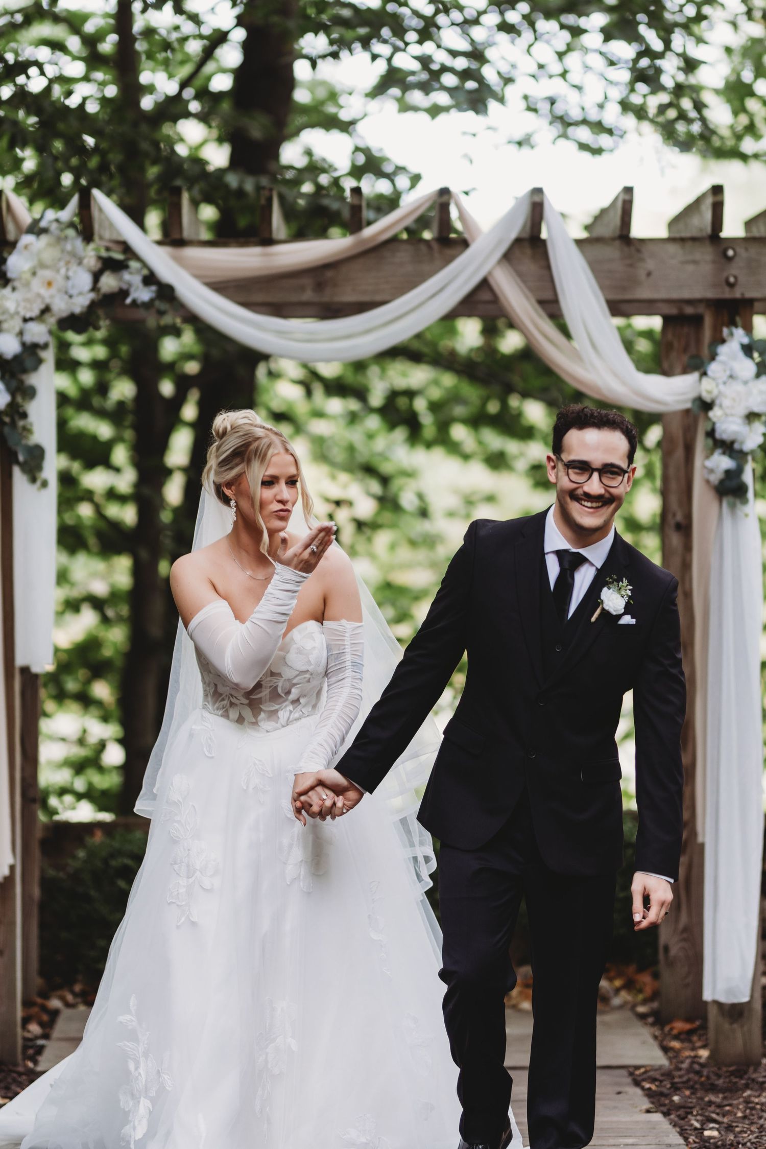 A joyful wedding couple shares a playful moment together under an outdoor ceremony arch decorated with white fabric.