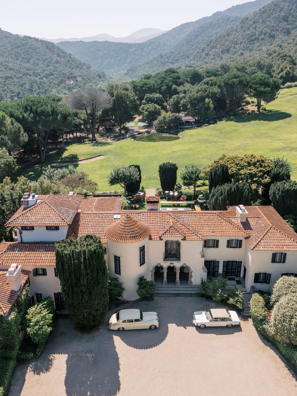 Aerial view of Spanish-style villa with terracotta roofs nestled in green mountains with multiple connected buildings.