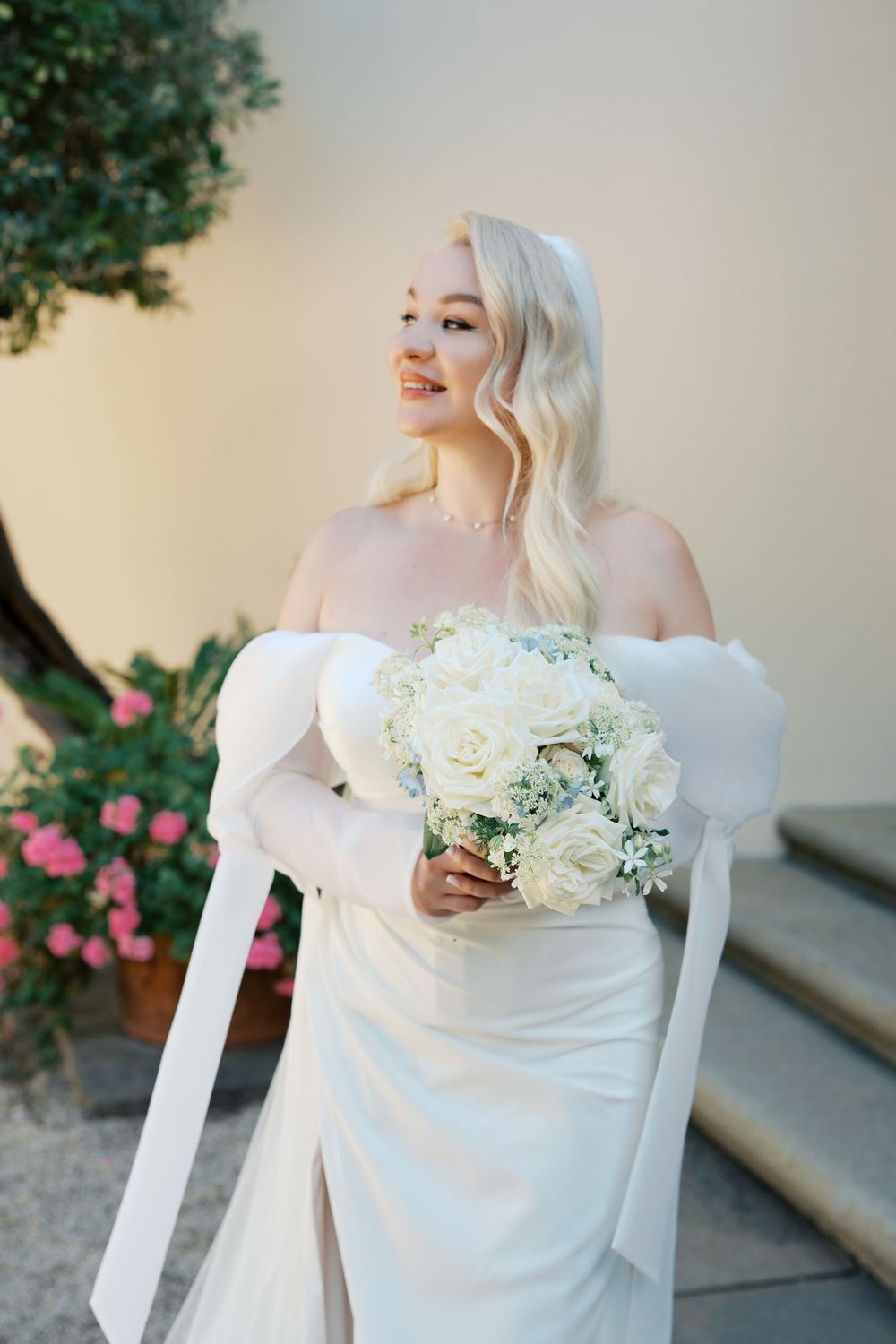A bride in an elegant off-shoulder white wedding dress holds a delicate bouquet of white roses and baby's breath.