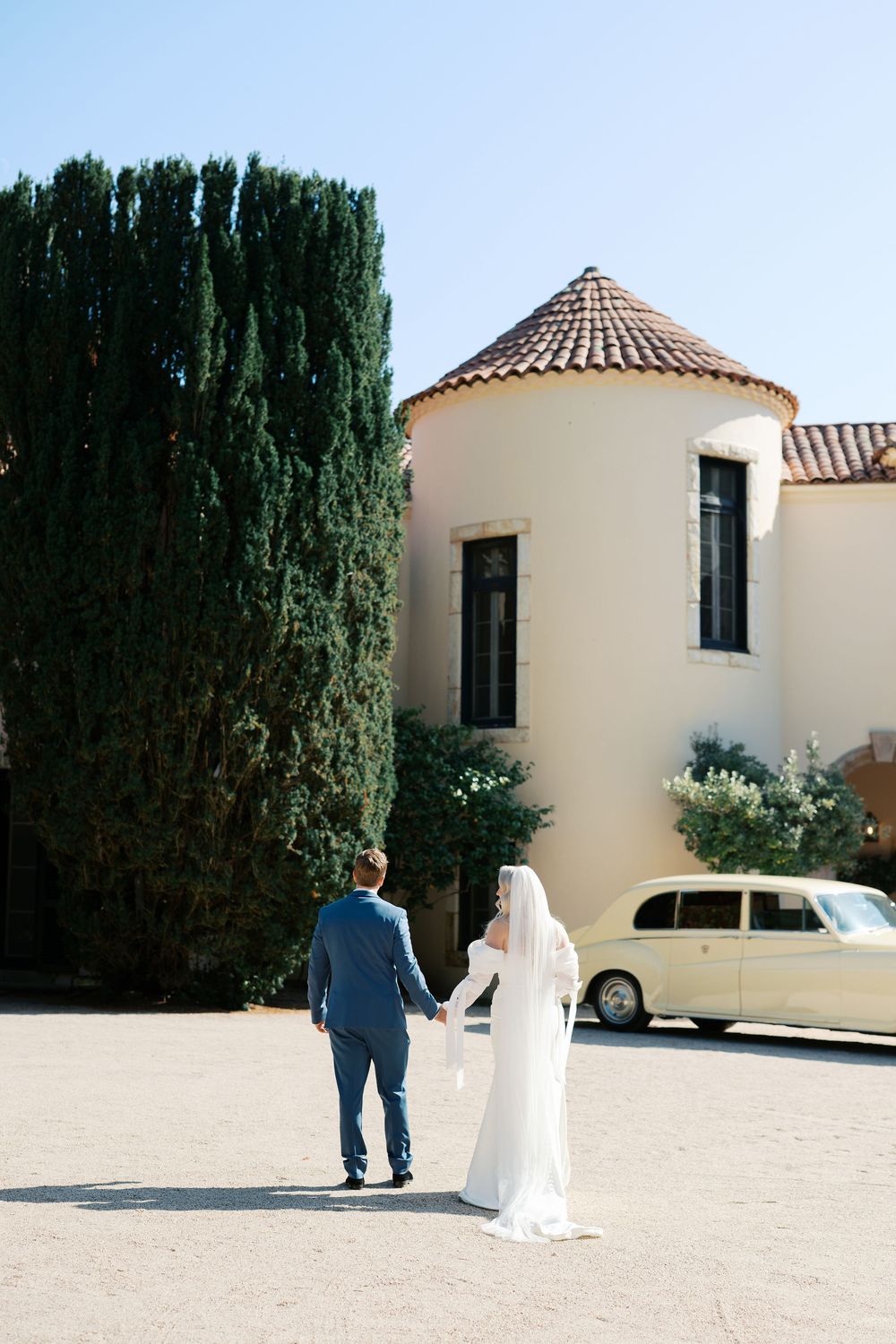 A couple walks hand in hand outside a Mediterranean-style villa with a vintage cream-colored car in the background.
