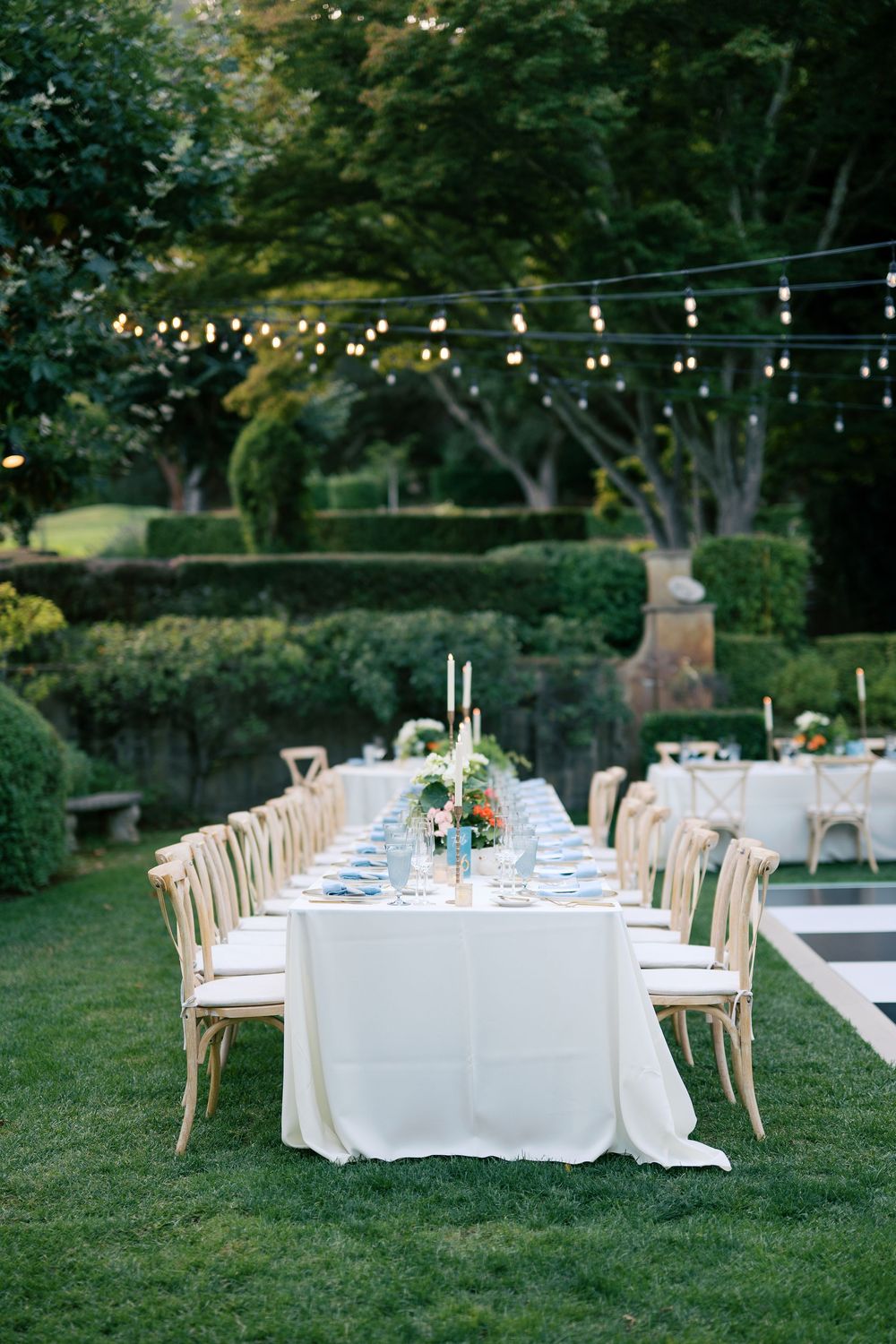 Elegant outdoor wedding reception setup with long tables, white linens and wooden chairs under string lights in a garden setting.