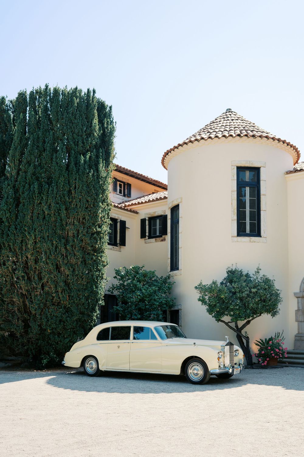 A classic cream-colored vintage car parked outside a Mediterranean villa with tall cypress trees.