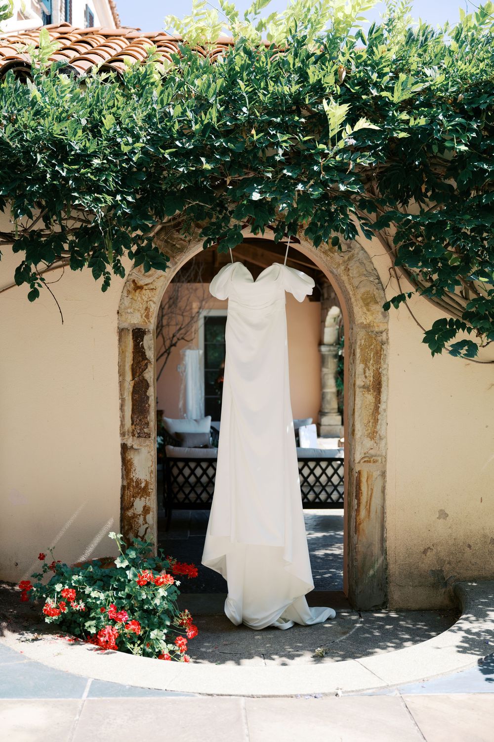 White wedding dress hanging from a rustic stone archway adorned with climbing vines and red flowers.