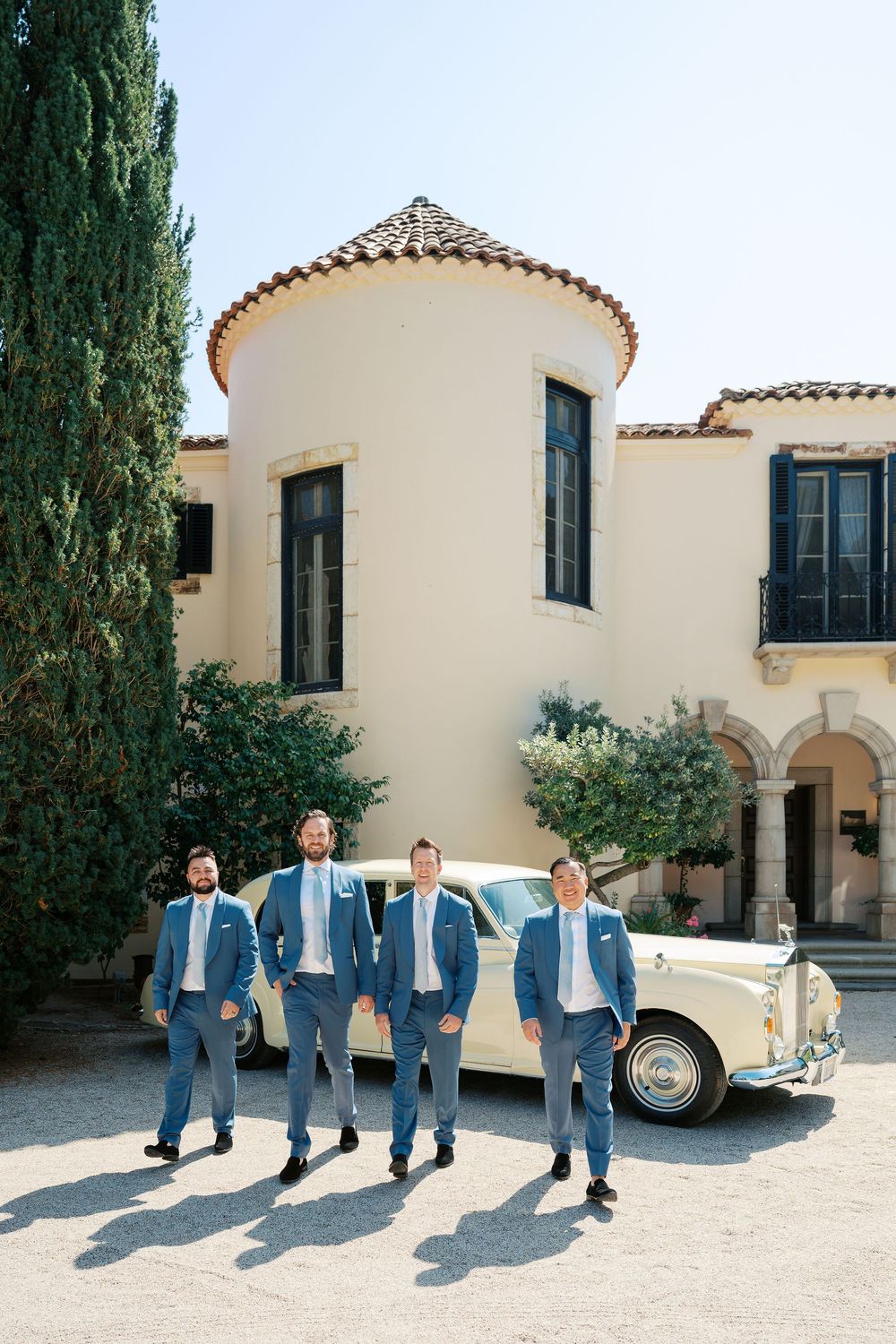 Groomsmen in blue suits pose with a vintage cream car in front of a Mediterranean villa.