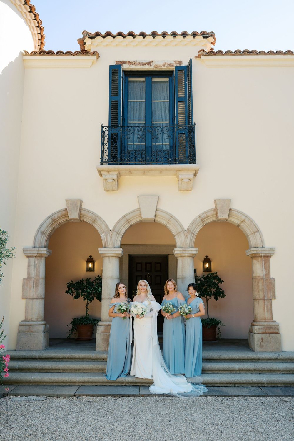 Bridesmaids in light blue dresses stand with the bride in white under a Mediterranean-style archway at a wedding venue.