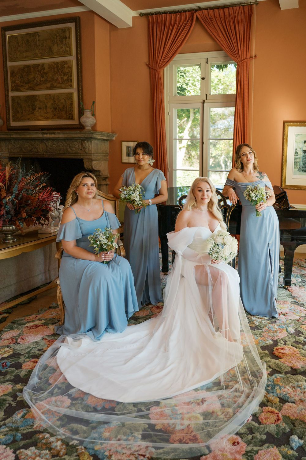Bridesmaids in light blue dresses help arrange the bride's flowing white wedding gown train on a floral carpet.