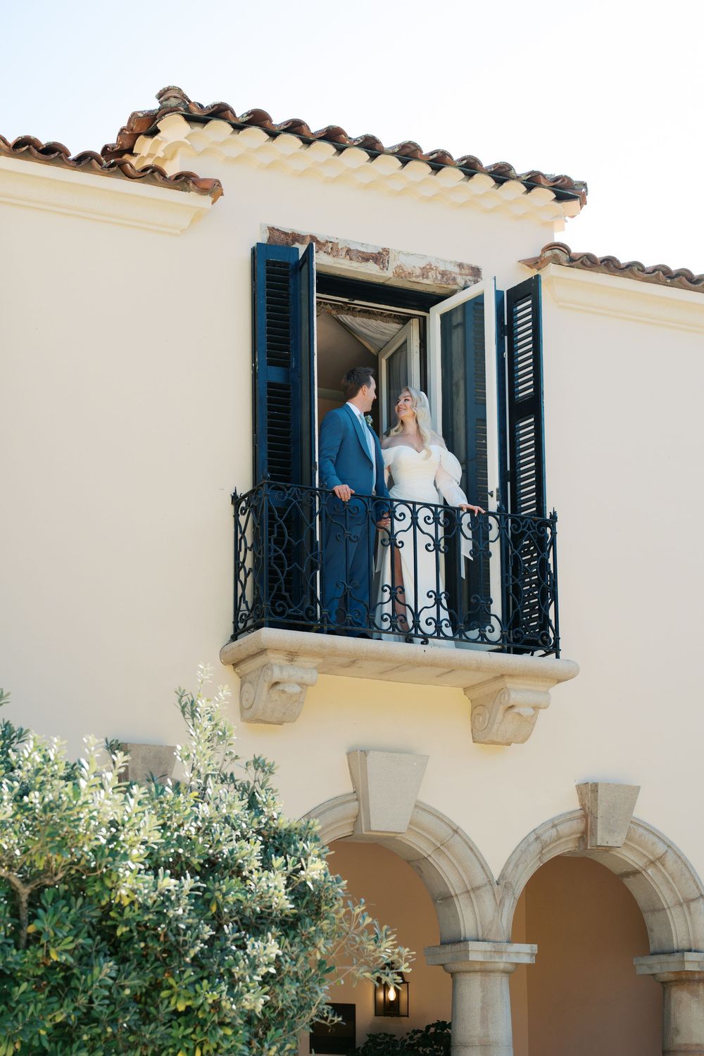 Spanish colonial balcony with dark blue shutters and ornate ironwork details against white stucco facade.