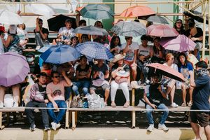 Spectators hold colorful umbrellas while watching an outdoor event on a sunny day.