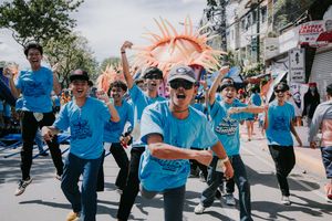Energetic performers in light blue shirts dancing and celebrating during an outdoor street parade.