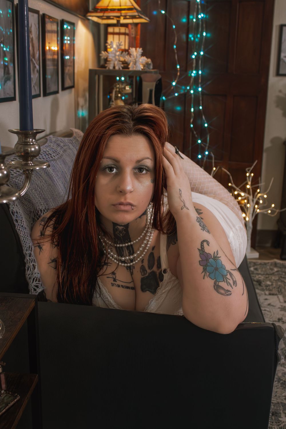 Tattooed redhead woman with pearl necklace sits on couch near twinkling blue fairy lights.